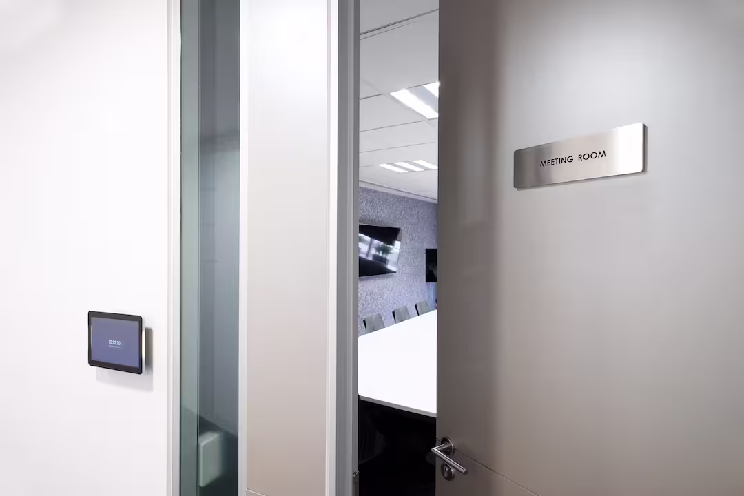 Eine Innenansicht des Eingangs zu einem Besprechungsraum. Eine leicht geöffnete Tür mit einem silbernen Schild mit der Aufschrift "MEETING ROOM" in Großbuchstaben und einer Türklinke ist auf der rechten Seite zu sehen. Links an der weißen Wand hängt ein Tablet. Im Hintergrund sind ein langer weißer Konferenztisch, Stühle und eine strukturierte graue Wand im Raum sichtbar. Die Decke hat Einbauleuchten.