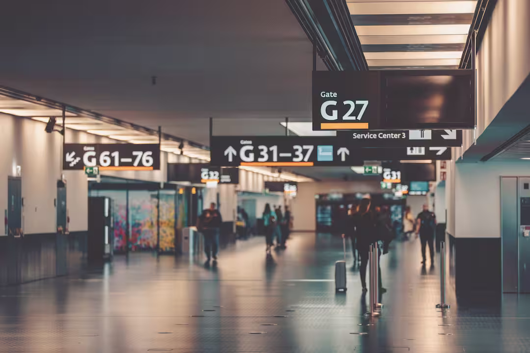 Ein Blick einen langen Flughafen-Terminalgang hinunter mit klaren schwarzen Hängeschildern. Ein großes Schild im Vordergrund zeigt "Gate G 27" und "Service Center 3" an. Weitere Schilder weiter hinten weisen zu "↑ Gates G 31-37" und "← Gates G 61-76". Menschen mit Koffern gehen den Gang entlang, und die Lichter spiegeln sich auf dem glänzenden Boden. Der Hintergrund ist leicht unscharf.