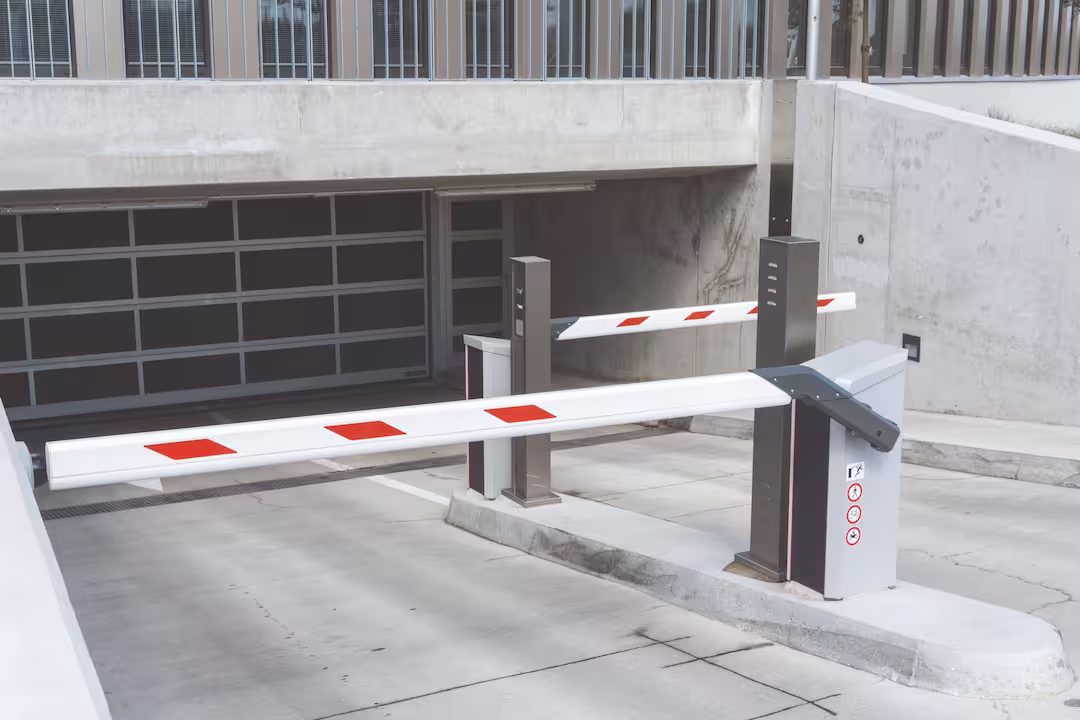 Eine Nahaufnahme einer Tiefgarageneinfahrt mit zwei geschlossenen rot-weiß gestreiften Schranken. Die vordere Schranke steht rechts, die hintere Schranke links. Im Hintergrund ist ein geschlossenes Rolltor und Betonwände zu sehen. Schilder mit Verbot für Fußgänger und Radfahrer sind an der vorderen Schrankensäule befestigt. Der Boden besteht aus Betonplatten.